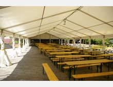 Großes Festzelt mit gelben Holzbänken und Tischen in Reihen auf einem gepflasterten Boden. Das Zelt ist mit weißen Planen und Querstreben überdacht, die Seiten sind offen und bieten Blick auf Bäume und eine Backsteinmauer im Hintergrund.
