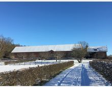 Ländliche Winterlandschaft mit einem langen, schneebedeckten Scheunengebäude aus Holz und einem Weg, der links und rechts von niedrigen Hecken gesäumt ist. Der Himmel ist klar und blau, Bäume sind im Hintergrund erkennbar.