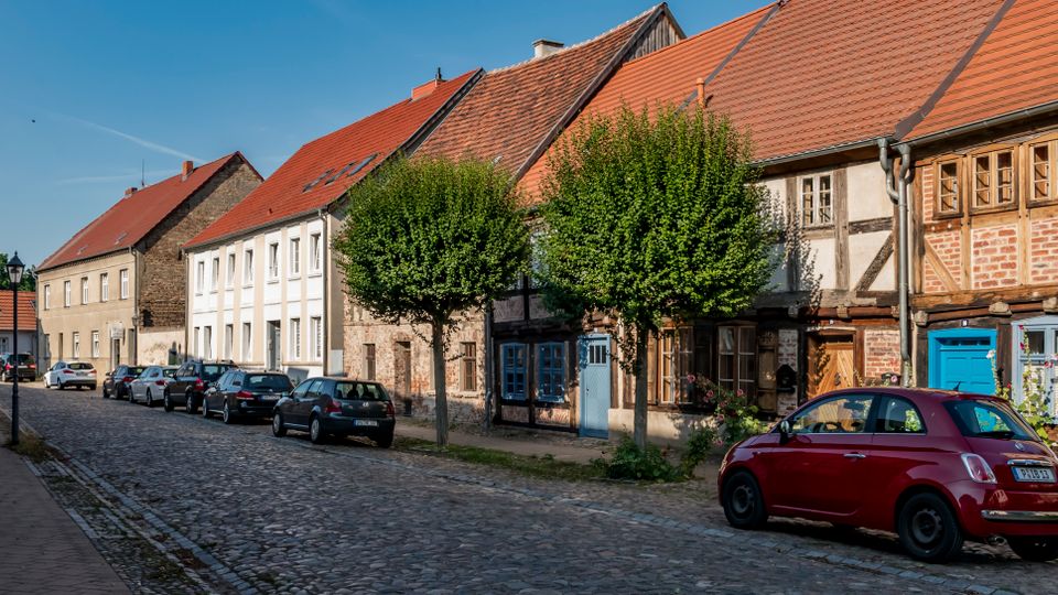 Kopfsteingepflasterte Straße in einer Altstadt mit parkenden Autos, gesäumt von niedrigen Wohnhäusern mit roten Ziegeldächern und teils sichtbarem Fachwerk. Entlang der Häuser stehen gleichmäßig geschnittene Bäume, im Hintergrund verläuft die Straße leicht nach links.