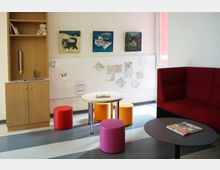Ein heller Aufenthaltsraum mit niedrigen, bunten Hockern und runden Tischen. An der Wand hängen Bilder und Karten, daneben steht ein Holzschrank mit Regal und eingebautem Waschbecken; rechts befindet sich eine rote, gepolsterte Sitzbank.