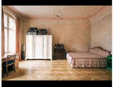 Ein schlicht eingerichtetes Zimmer mit einer beigefarbenen Wand, bestehend aus einem Holzboden, einem weißen Kleiderschrank mit dekorativen Details, einem Einzelbett mit floralem Muster und einem Schreibtisch mit Stuhl. Alte Koffer sind auf dem Schrank und einer Kommode gestapelt, und ein großes Fenster mit orangen Vorhängen lässt Tageslicht herein.