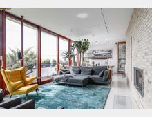 Modernes Wohnzimmer mit einer großen Fensterfront, die auf eine Terrasse mit Pflanzen blickt. Im Raum befinden sich ein graues Ecksofa, ein gelber Sessel, eine Ziegelwand mit integriertem Kamin, ein türkisfarbener Teppich sowie mehrere Pflanzen und dekorative Objekte.