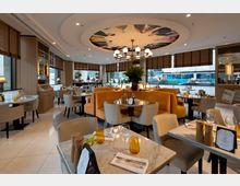 Ein stilvoll eingerichtetes Restaurant mit hellen Möbeln, Marmortischen und gepolsterten Sitzbänken in orangefarbenem Stoff. Große Fenster mit Holzjalousien bieten Ausblick nach draußen, eine zentrale Deckenleuchte und eine Deckenmalerei verleihen dem Raum Eleganz.