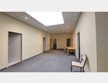 Ein heller, rechteckiger Raum mit beigem Anstrich, grauem Teppichboden und einer Rasterdecke mit eingebauten Leuchten. Der Raum hat mehrere Türen, die in angrenzende Bereiche führen, sowie einfache Möbel, darunter einen Tisch und einen Stuhl.