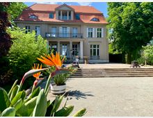 Zweistöckige Villa mit hellem Putz, rotem Ziegeldach und symmetrischen Fenstern, inmitten eines Gartens mit Blumen und Bäumen; im Vordergrund leuchtend orangefarbene Paradiesvogelblumen in einem Topf und eine breite Steintreppe, die zur Terrasse des Hauses führt.