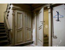 Treppenhaus in einem alten Gebäude mit abgenutzten Holztüren und Geländern, sowie abgeplatztem Putz an den Wänden. Eine geöffnete Tür führt in einen schwach beleuchteten Raum, an der Wand hängt ein Kleiderbügel.