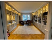 Wohnzimmer mit Holzparkettboden, großen weißen Einbauregalen an den Wänden, die Fotos, Bücher und dekorative Objekte enthalten, sowie einem Flügel in der Mitte. Ein großes Fenster mit Vorhängen im Hintergrund lässt Tageslicht herein.