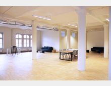 Großer, heller Innenraum mit Holzboden, hohen Decken und weißen Säulen. Der Raum ist mit einer Kombination aus Sofas, Tischen und Stühlen ausgestattet; große Fenster mit Rundbögen lassen viel Licht herein.