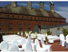 Eine Dachterrasse mit roten Backsteinen, auf der Stühle und Bänke mit weißen Hussen für eine Veranstaltung, vermutlich eine Hochzeit, aufgestellt sind; dekoriert mit weißen Blumen in hohen Glasvasen. Im Hintergrund befindet sich ein großes historisches Gebäude mit roter Ziegelfassade und markanten Schornsteinen.