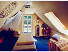 Ein gemütlicher Behandlungsraum unter einer Dachschräge, mit einem Massagetisch in der Mitte, einem Oberlicht rechts und einer Glastür im Hintergrund. Der Raum ist mit einem kleinen Holztisch, einer Kommode, Pflanzen, dekorativen Gegenständen und einem Mandala-Muster an der Wand ausgestattet.