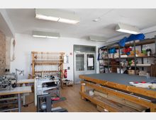 Werkstatt mit Holz- und Näharbeitsgeräten. Ein großer Arbeitstisch, Regale mit Materialien, Nähmaschinen und ein Webstuhl prägen den Raum. Die Wände sind teils aus weißem Putz, teils aus sichtbarem Ziegelstein, und an der Decke befinden sich große Leuchtstofflampen.