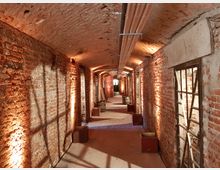 Langer, schmaler Ziegelsteinkeller mit gewölbter Decke, rustikalen Backsteinwänden und beleuchteten Wandleuchten. Der Raum ist spärlich ausgestattet mit Metallrohren an der Decke, einigen Pflanzkübeln und Holzfässern.