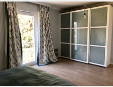 Schlafzimmer mit einem großen weißen Kleiderschrank mit Milchglasfronten an der rechten Wand, einem bodentiefen Fenster mit floral gemusterten Vorhängen links, Holzboden und einer grünen Bettdecke im Vordergrund.
