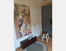 Innenraum einer Wohnung mit einer hellen Wand, an der ein großes, farbenfrohes Gemälde hängt. Rechts steht ein grauer Einbauschrank mit einem weißen Hocker davor, am Boden liegt ein runder Teppich aus hellem Material, und im Vordergrund befindet sich eine dunkle Holztruhe.