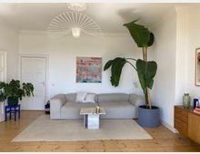Bright, minimalist living room with white walls, wood floors, and a large gray sectional sofa on a light area rug. A small marble-topped coffee table sits in front of the sofa, with a large potted plant, wall art, and a sculptural ceiling light above.