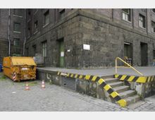 Ehemaliges Industriegebäude aus dunklem Ziegel- und Steinmauerwerk mit großen grünen Türen und Fenstern. Im Vordergrund befinden sich eine gelbe Metalltreppe mit schwarz-gelben Warnmarkierungen, ein orangefarbener Abfallcontainer und einige Verkehrshütchen.