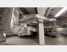 Industriegebäude mit einem technischen Raum, in dem große Lüftungsrohre und Leitungen verlaufen, die mit silberner Isolierung ummantelt sind. Die Umgebung ist funktional gestaltet, mit sichtbaren Betonpfeilern, hellen Wänden und einer Beleuchtung durch Neonröhren.