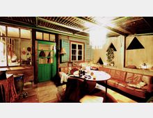 Ein rustikaler Innenraum mit einer Sitzgruppe um einen runden Tisch, einer braunen Lederbank und verschiedenen dekorativen Elementen. Die Wände aus Holz und Putz haben dreieckige Fenster, und hinter einer grünen Tür befinden sich weitere Räume.