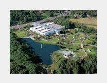 Luftaufnahme eines weitläufigen Geländes mit einem See in der Mitte, umgeben von Grünflächen, Bäumen und Wegen. Im Hintergrund sind große, rechteckige Gebäude und landwirtschaftliche Felder zu sehen.