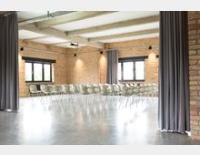 Ein moderner Seminarraum mit Wänden aus Sichtziegeln, einem glänzenden Betonboden und großen Fenstern mit schwarzen Rahmen. Reihen aus hellgrauen, stapelbaren Stühlen sind aufgestellt, und graue Vorhänge teilen den Raum teilweise ab.