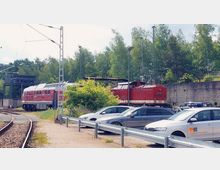 Ein kleiner Bahnhof oder Werkstattbereich mit zwei roten Diesellokomotiven, einer Schiene im Vordergrund und geparkten Autos daneben. Im Hintergrund befindet sich eine Betonwand, dahinter sind Bäume sichtbar.