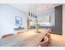 Heller, modern eingerichteter Ess- und Küchenbereich mit langem Holztisch und gepolsterten Stühlen, darüber mehrere zylindrische Pendelleuchten. Im Hintergrund eine minimalistische Einbauküche mit grauen Fronten, hoher Schrankwand, Arbeitsflächenbeleuchtung und Holzfußboden.