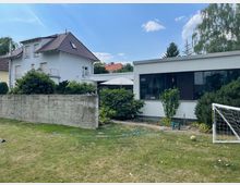 Ein Garten mit einer Rasenfläche, auf der ein kleines Fußballtor und ein Ball liegen, umgeben von Hecken und Sträuchern. Im Hintergrund sind zwei Gebäude zu sehen: links ein zweistöckiges Haus mit Satteldach, rechts ein flaches Gebäude mit großen Fenstern.