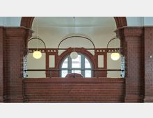 Innenraum mit rotbraunen Backsteinbögen und gemauerten Brüstungen, davor eine Galerie mit schwarzem Geländer. In der Mitte befindet sich ein großes Rundbogenfenster, flankiert von kleineren Bögen, mit drei hängenden Kugelleuchten vor einer hell verputzten Decke.