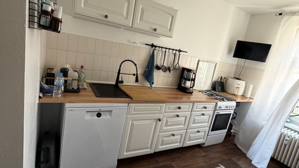 Schmale Küchenzeile in einem hellen Raum mit weißen Fronten, Holzarbeitsplatte und Fliesenspiegel; ausgestattet mit Spüle, Geschirrspüler, Herd/Backofen sowie Kaffeemaschine und Kleingeräten. Dunkler Holzboden mit gemustertem Teppich, Hängelampe und Fenster mit transparenten Vorhängen am rechten Rand.