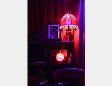 Ein Regal in einem gemütlich beleuchteten Raum mit roten Vorhängen, darauf eine Tischlampe mit rotem Schirm und Quasten, daneben eine Glasflasche und ein gerahmtes Foto. Im Regal befinden sich Bücher und eine leuchtende Kugel mit ringförmiger Struktur.