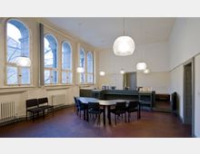 Innenraum eines Bürogebäudes mit hohen, rundbogigen Fenstern, die auf eine Außenterrasse blicken. Der Raum enthält einen rechteckigen Tisch mit schwarzen Stühlen, eine Empfangstheke und mehrere hängende Pendelleuchten sowie helle Wände und einen roten Boden.