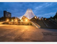 Architektonisches Gebäude mit markantem, weißem Zeltdach und angestrahlten Fassaden bei Dämmerung, umgeben von Treppen und einer großen Freifläche. Im Hintergrund steht ein modernes Hochhaus.
