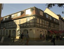 Fachwerkhaus an einer Straßenecke mit gelblicher Fassade und roten Dachziegeln, großen Fenstern und Rundbögen im Erdgeschoss. Menschen gehen auf dem Kopfsteinpflaster, rechts befindet sich ein Café mit roten Sonnenschirmen.