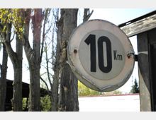 Ein altes Metallschild mit der Aufschrift „10 Km“ hängt an einem Pfosten, umgeben von hohen Bäumen mit teilweise blättrigen Ästen. Im Hintergrund sind ein dunkles Holzdach und eine hellere Gebäudeoberfläche sichtbar.