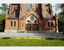Backstein-Kirchfassade mit gotischen Spitzbögen, großen Holztüren, einem runden Fenster und detaillierten Verzierungen; im Vordergrund ein gepflegter Garten mit bunten Blumenbeeten.