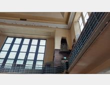 Innenansicht eines historischen Gebäudes mit hohen, vergitterten Fenstern, gelblichem Putz an den Wänden und dekorativen Metallgeländern. Auf der rechten Seite ist ein kleiner Balkon mit Ziegelsteinverkleidung sichtbar.