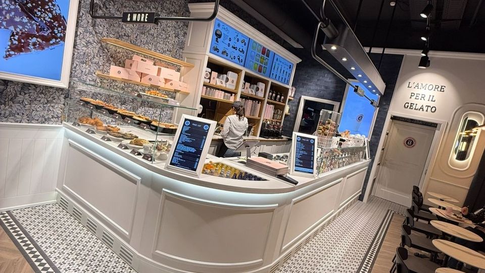 Moderner Eisladen mit geschwungener, weißer Verkaufstheke, darauf Speisen und digitale Displays. Der Raum hat dekorative Wandmuster, eine dunkle Decke mit Hängelampen, gemusterte Fliesenböden und eine Sitzbereich mit runden Tischen und Stühlen.