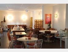 Ein gemütlicher Innenraum eines Cafés mit verschiedenen Tischen und Stühlen, überwiegend aus Holz und Metall. An der Wand hängen runde Wandlampen, ein buntes Poster und ein Kaktus steht in einer Ecke; links ist eine gelbe Gardine sichtbar.