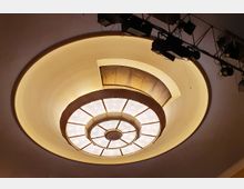 Kreisförmige Deckenleuchte mit einem konzentrischen Design aus Glas- und Metallteilen, eingebettet in eine Deckennische mit warmem indirektem Licht. Im oberen Bereich sind Bühnenscheinwerfer und technische Träger zu erkennen.