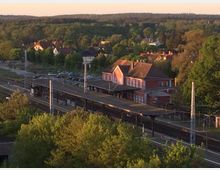 Bahnhof in einer Kleinstadt mit einem zweistöckigen, roten Hauptgebäude und mehreren Bahnsteigen. Im Vordergrund befinden sich Bäume, während im Hintergrund parkende Autos, Wohnhäuser und ein Wald zu sehen sind.
