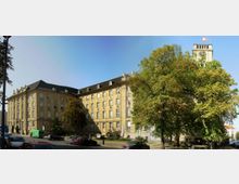 Historisches Verwaltungsgebäude aus gelbem Stein mit mehreren Etagen und einem dunklen Schrägdach, flankiert von einem Turm mit Uhr und Fahne. Im Vordergrund stehen große Bäume, Autos auf einer Straße und ein Gehweg.