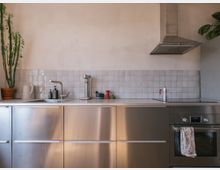 Moderne Küche mit einer Arbeitsplatte aus weißem Material, einem Spülbecken, Küchengeräten und einem Herd mit Edelstahlhaube. Die unteren Schränke sind aus gebürstetem Edelstahl, die Rückwand ist mit kleinen weißen Fliesen verkleidet, an der linken Seite steht ein hoher Kaktus in einem Topf.