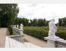 Steinbalustrade mit weißen Marmorstatuen und Vasen, umgeben von gepflegten Hecken und Wegen in einer parkähnlichen Landschaft. Im Hintergrund sind große Bäume und ein wolkiger Himmel zu sehen.