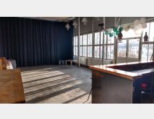 Leerer Veranstaltungsraum mit großen Sprossenfenstern, schwarzem Vorhang, einer Holztheke im Vordergrund und vereinzelten Luftballons an der Decke. Der Betonboden reflektiert das einfallende Tageslicht durch die Fenster.