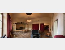 Ein historischer Veranstaltungsraum mit einer kleinen Bühne im Hintergrund, dekoriert mit einer bogenförmigen Bordüre. Der Raum hat hohe Decken mit einem Kronleuchter, große Fenster mit roten Vorhängen und Holzböden mit Stuhl- und Tischanordnung.