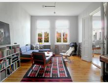 Helles Wohnzimmer mit Holzboden und modernen Möbeln, darunter ein großes graues Ecksofa, ein Sessel, ein Bücherregal und ein bunter Teppich. Durch zwei große Fenster ist eine Aussicht auf Dächer und Bäume sichtbar, rechts führt eine geöffnete Tür in ein weiteres Zimmer.