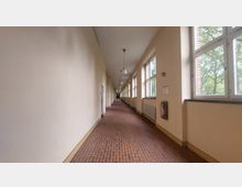 Langer, heller Flur in einem Gebäude mit cremefarbenen Wänden, rotbraunem Ziegelboden und einer Reihe großer Fenster auf der rechten Seite, die Blick auf Bäume gewähren. An der Wand befinden sich Heizkörper, eine Feuermeldeeinrichtung und Hängelampen an der Decke.