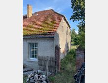 Ein altes, verputztes Wohnhaus mit schrägem Dach aus roten Ziegeln, teilweise mit Moos bewachsen. Ein kleines Fenster mit weißen Rahmen ist an der Vorderseite sichtbar, daneben ein einfacher Holzzaun und zwei rote Backsteinpfeiler. Im Hintergrund befindet sich eine grüne, ländliche Umgebung mit Bäumen.