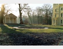 Eine parkähnliche Landschaft mit einem kleinen, runden Brunnen in der Mitte, umgeben von Rasenflächen und kahlen Bäumen. Links steht ein pavillonartiges Gebäude mit Säulen, rechts ist ein heller, klassizistischer Gebäudeteil sichtbar.