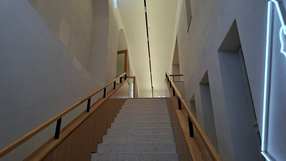 Blick von unten auf eine breite Innentreppe mit hellen Stein-Stufen und Holzgeländern, die zu einem schmalen, hohen Flur führt. Weiße Wände mit Nischen und Bögen sowie eine lange Deckenleuchte prägen den modernen Innenraum.
